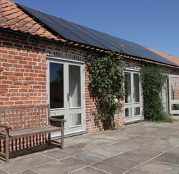 A brick building with white framed glass doors and solar panels on the roof with a wooden bench outside at Cow Byre in Malton