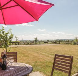 A garden with a wooden table and chairs under a pink umbrella and cows grazing in the field at Cow Byre in Malton