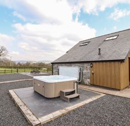 An outdoor hot tub on a tiled platform next to a stone and wood building with a fenced field in the background at Beudy Bach Barn in Llanuwchllyn near Bala