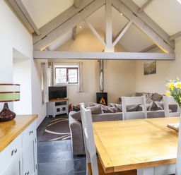 A dining table with chairs and flowers in a room with exposed beams a wood stove a sofa and a television at Beudy Bach Barn in Llanuwchllyn near Bala