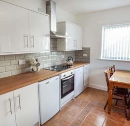 A kitchen with white cabinets wooden countertops a stove with an oven a kettle a small fridge a wooden dining table with four chairs and a window with vertical blinds at Arfryn in Bethesda