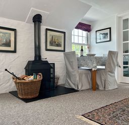A dining area with a small table and two covered chairs next to a wood-burning stove and basket with firewood at The Boat House in Golant near Tywardreath