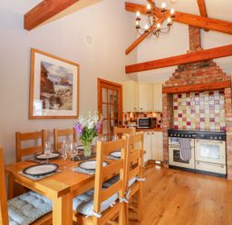 A kitchen with wooden dining table and chairs set with plates and glasses a stove with brick surround and tiled backsplash and wooden beams on ceiling at The Wagon House Tresmeer near Launceston
