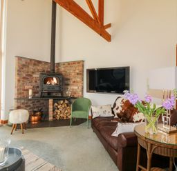 A living room with a wood burning stove and stacked firewood a leather sofa a green chair a wicker side table with flowers and a lamp a wall mounted television at The Wagon House in Tresmeer near Launceston