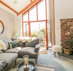 A living room with a gray sectional sofa a round window a lit fireplace and large wooden framed glass doors at The Wagon House in Tresmeer near Launceston