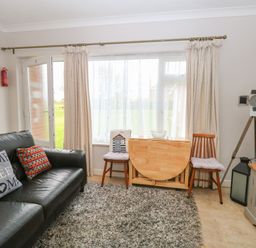 A living room with a black leather sofa and cushions a wooden folding table two chairs a large window with curtains a gray carpet a tripod lamp and a television unit at En-Casa in Mundesley