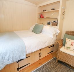 A small bedroom with a bed against the wall a wicker chair and built in shelves at Shepherds Hut in Castleton Peak District