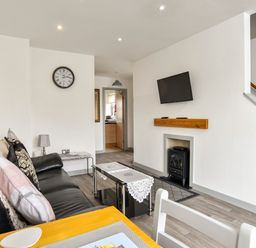 A living room with a black leather sofa cushions a wall clock a wall mounted TV and a small stove at Plas Bach in Aberffraw