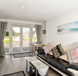 A living room with a black and white sofa with cushions a glass coffee table a wall-mounted television and double glass doors at Plas Bach in Aberffraw