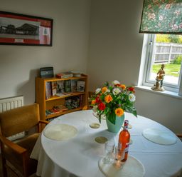 A dining room with a round table covered with a white tablecloth flowers in a vase on the table wooden chairs a window with floral blinds a small bookshelf with books and a figurine on the windowsill at Anvil Lodge in Goulceby near Louth