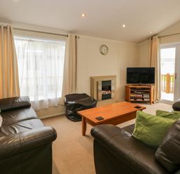A living room with brown leather sofas a wooden coffee table a wall clock a tv on a wooden stand and cream curtains at Jolie in Hunstanton