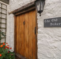 A wooden door with a black lantern and a sign that says The Old Bakery on a white textured wall with red and orange flowers nearby at The Old Bakery in Crowcombe