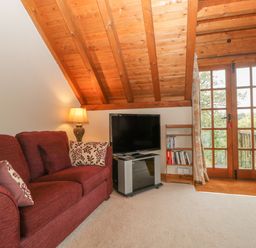 A living room with a red sofa, a table lamp, a television on a stand, a small bookshelf with books, and glass doors with floral curtains at Floras Barn in Crowcombe