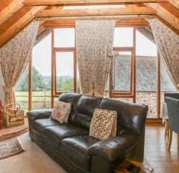 A living room with a black leather sofa a red wood-burning stove floral curtains and a dining table with chairs at Floras Barn in Crowcombe