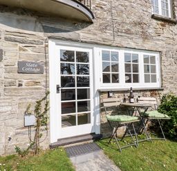 A stone cottage exterior with a white framed glass door and windows a small table with two chairs and cushions on grass at Slate Cottage Tredethy near Bodmin Moor