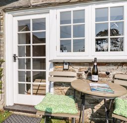 A small outdoor table with two chairs and a bottle of prosecco outside a stone cottage with white framed door and windows at Slate Cottage Tredethy near Bodmin Moor