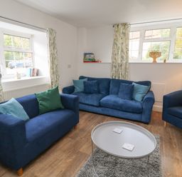 A living room with blue sofas green cushions a round gray table and windows with patterned curtains at Aldrich Cottage in Radstock