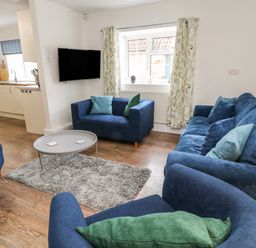 A living room with blue sofas and a round gray table on a rug with a kitchen area in the background at Aldrich Cottage in Radstock