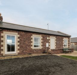 A single-story brick house with three windows and a door with a small patio area with benches at West Gate in Embleton