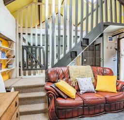 A sitting area with a brown leather sofa with yellow pillows and stairs with railing in a cottage at Celyn Farm Cottage Deiniolen near Llanberis