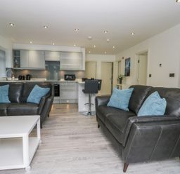 A living room with two black leather sofas and blue pillows a white coffee table and kitchen area in the background at The Office Gardens Apartment 2 in Whitby