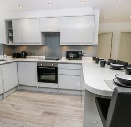 A kitchen with grey cabinets a built-in oven a microwave toaster kettle and a counter with black chairs and place settings at The Office Gardens Apartment 2 Whitby