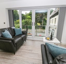 A living room with black leather sofas and blue cushions facing glass doors overlooking greenery at The Office Gardens Apartment 2 in Whitby