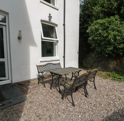 An outdoor seating area with a metal table and benches on gravel near a white building and greenery at Dale Cottage in Llanrwst