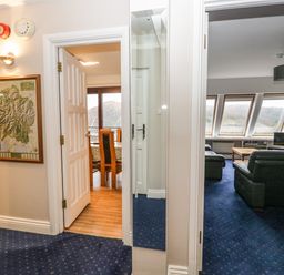 A hallway with blue carpet and open doors leading to a dining area with dining table and chairs and a living room with sofas and windows at Rothay 17 in Ambleside