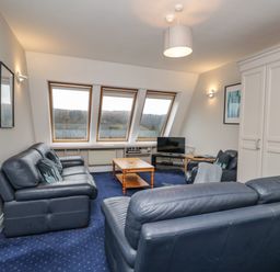 A living room with blue leather sofas a wooden coffee table a television and three large windows at Rothay 17 Ambleside