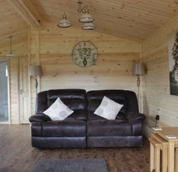 A living room with a dark sofa two pillows a clock wall lamps wooden tables with flowers and a tree picture at Hayfields in Grimston