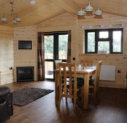 A wooden dining table with chairs in a wooden cabin room with a kitchen area a sofa and a wall-mounted television at Hayfields in Grimston