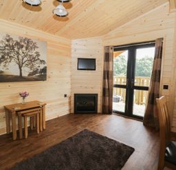 A wooden cabin room with nested tables a chair a wall-mounted TV a fireplace and glass doors leading outside at Hayfields in Grimston