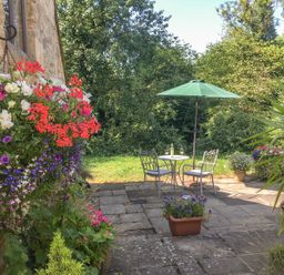 A garden patio with a small round table and two chairs under a green umbrella surrounded by green plants and colorful flowers at The Old Cider House in Prestbury near Cheltenham