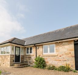 The front exterior of a single-story stone house with a black door, windows, a garage door, and a paved driveway with plants along the side at The Croft in Longhoughton near Alnwick