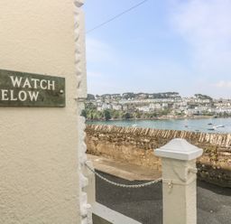 A street view with a wall and a sign that reads The Watch Below overlooking a body of water and houses on a hillside at The Watch Below in Polruan
