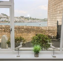 A window with two bird figurines and a small plant on the sill overlooking a stone wall a water body with boats and houses in the background at The Watch Below in Polruan