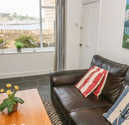 A living room with a dark leather sofa with striped cushions and a wooden table with flowers in a pot by a window showing a seaside town at The Watch Below in Polruan