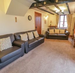 A living room with two brown leather sofas with floral cushions a wooden cabinet a window with curtains and a wall clock at Bank End Barn in Grizebeck