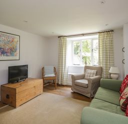 A living room with a green sofa with cushions a striped armchair a wooden chest used as a tv stand a wicker chair and curtains at Hall Bank Cottage in Rydal near Ambleside