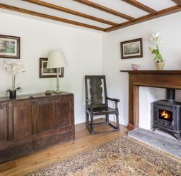 A corner of a room with a wooden chest, a lamp, a wooden chair, a fireplace with a wood stove, and framed pictures on the walls at Hall Bank Cottage in Rydal near Ambleside