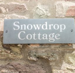 A stone wall with a grey rectangular sign reading Snowdrop Cottage in Laugharne