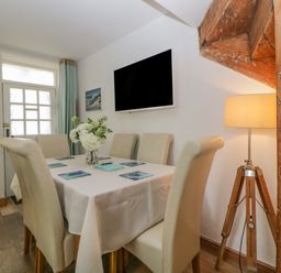 A dining area with six chairs around a table with a white tablecloth and a flower centerpiece wooden tripod lamp and wall mounted television at Curlews Cottage in Looe