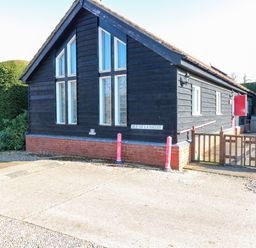 A black wooden chalet with large windows and a red brick base in a garden at Rookyard Cottage Holton near Halesworth