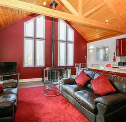 A living room with black leather sofas and red cushions a red carpet a glass coffee table a wood stove and a kitchen area in the background at Rookyard Cottage Holton near Halesworth