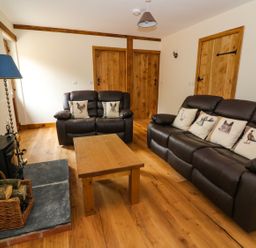 A living room with brown leather sofas with animal print cushions a wooden coffee table a wood stove with a basket of firewood and wooden doors at Ploony Hill Lodge in Bleddfa