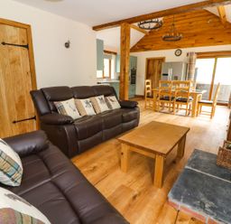 A living room with brown leather sofas, wooden coffee table, dining table with chairs, wood stove, and kitchen area at Ploony Hill Lodge in Bleddfa