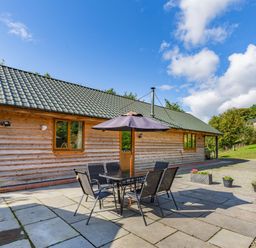 A wooden lodge with outdoor table and chairs under an umbrella on a paved patio at Ploony Hill Lodge in Bleddfa