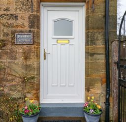 A white door with a mail slot and gold handle flanked by flower pots and a stone wall with a Jasmine Cottage sign