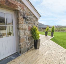 A stone building with a white door and outdoor plants on a wooden deck overlooking a grassy area at Horseshoe Cottage in Abersoch